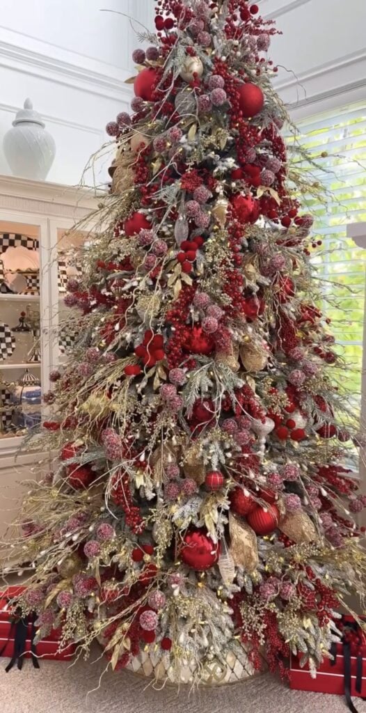 Glam Christmas tree in red and gold with cascading satin ribbons, oversized ornaments, glittered berry sprays, frosted foliage picks, and a patterned tree collar in a luxe living room.
