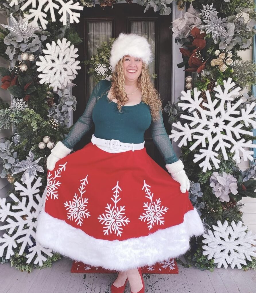 Katy from The Porch Club standing on a festively decorated Christmas porch, wearing a holiday outfit in front of layered winter greenery and ribbons