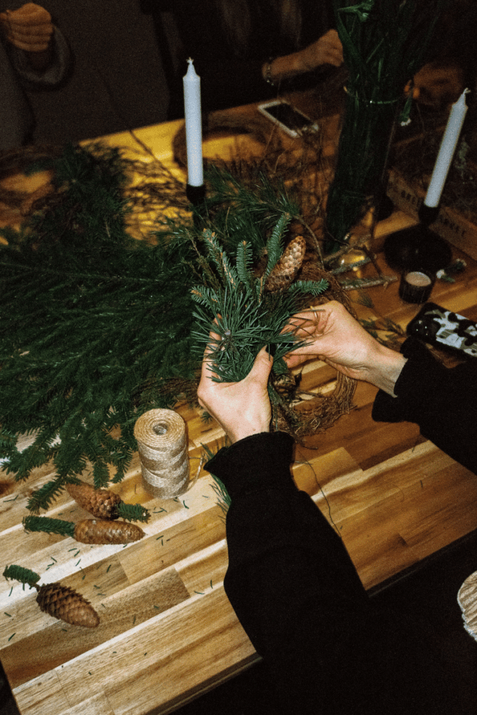 Image of greenery on a wooden table with a woman's hands holding greenery and creating a winter garland with a candle in the background
