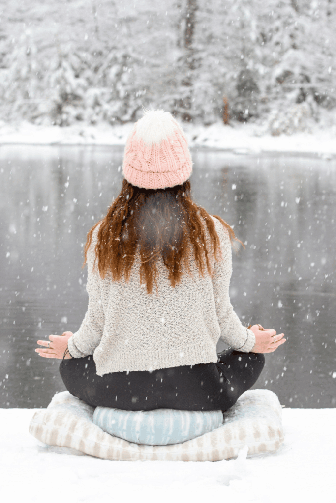 Image of the back of a girl sitting on two pillows in the snow meditating in front of a river as snowflakes fall