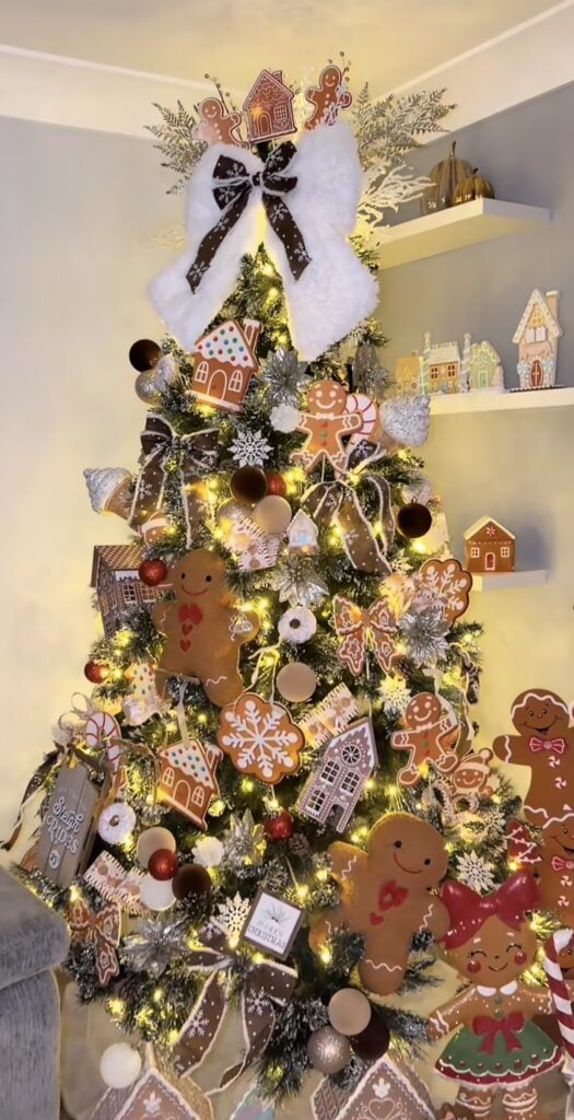 Gingerbread Christmas tree next to a glowing gingerbread house display, with cookie ornaments and soft white lights in a warm holiday corner.