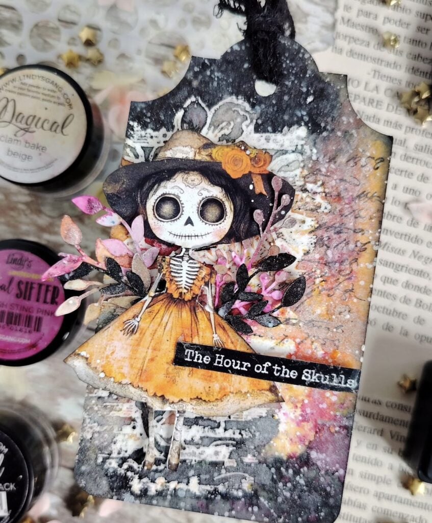 Mixed media tag featuring a skeleton doll in an orange dress with layered ephemera and splattered paint