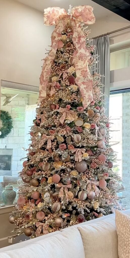 Tall Christmas tree densely decorated with blush pink and champagne ornaments, roses, berries, floral ribbon, and a large floral bow topper.