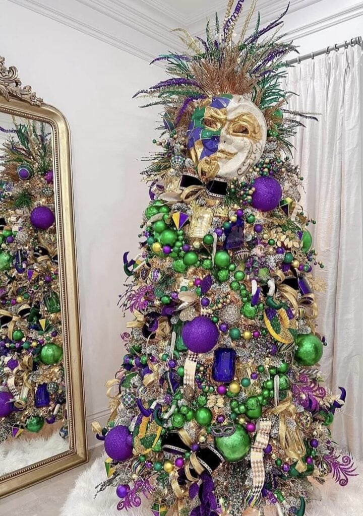 Glittery Mardi Gras tree with a large masquerade mask topper, packed with green and purple baubles, gold ribbon, and bead garlands, standing beside an ornate mirror.