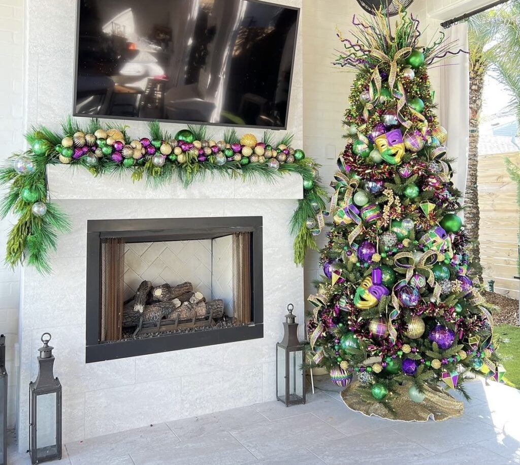 Outdoor patio Mardi Gras tree beside a modern fireplace, decorated with Mardi Gras masks, bead strands, and shiny ornaments in classic Mardi Gras colors, with a coordinating fireplace garland.