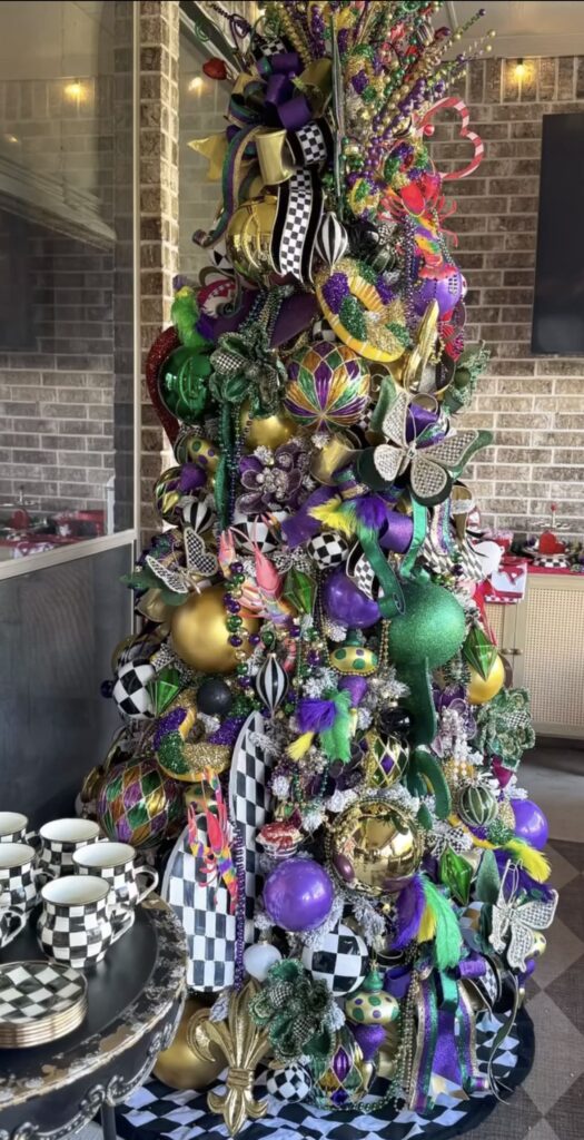 Outdoor patio Mardi Gras tree beside a modern fireplace, decorated with Mardi Gras masks, bead strands, and shiny ornaments in classic Mardi Gras colors, with a coordinating fireplace garland.