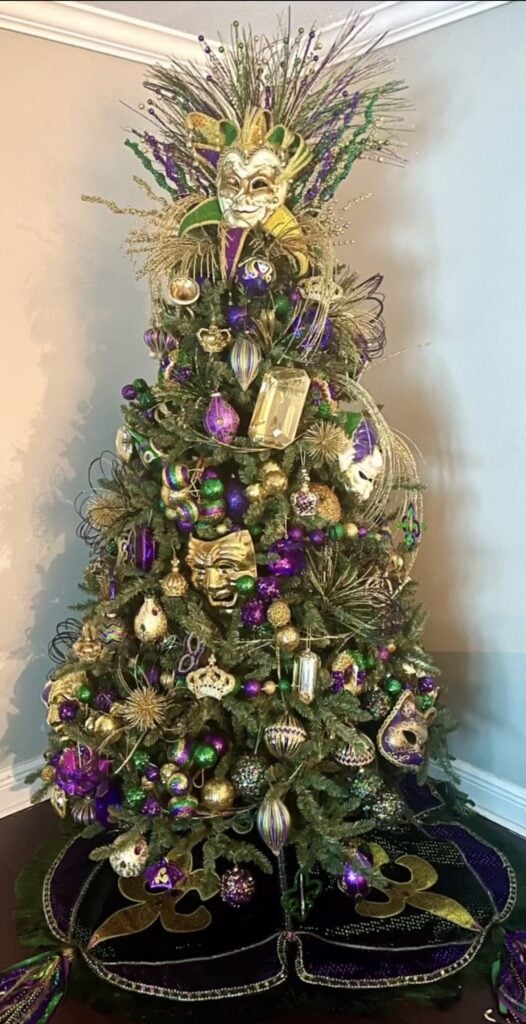Glittery Mardi Gras tree with a large masquerade mask topper, green and purple ornaments, gold ribbon, and bead garlands beside an ornate mirror.