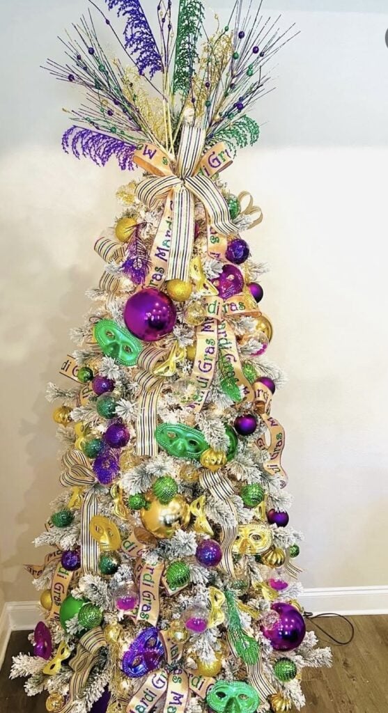 White Mardi Gras tree decorated with ribbon printed “Mardi Gras,” green mask ornaments, purple and gold baubles, and a tall sparkly spray topper.