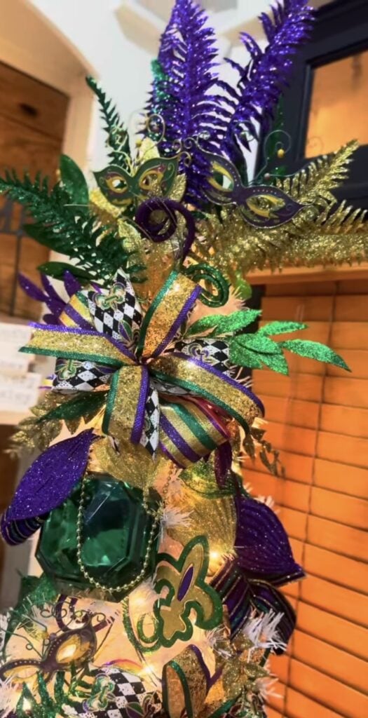 Mardi Gras tree topper with purple feather sprays, green and gold masquerade masks, and a purple, green, and gold ribbon bow.