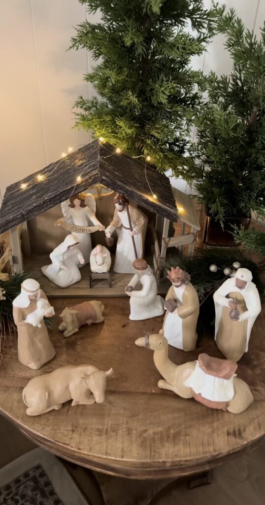 Cream and beige nativity figures arranged on a round wooden table beneath a rustic stable wrapped in warm white lights and surrounded by evergreen trees.