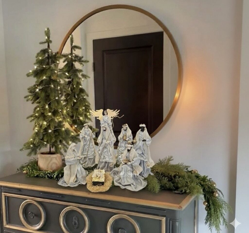 Elegant white and silver nativity figures displayed on a dark console with evergreen garland, a softly lit tabletop Christmas tree, and a round mirror.