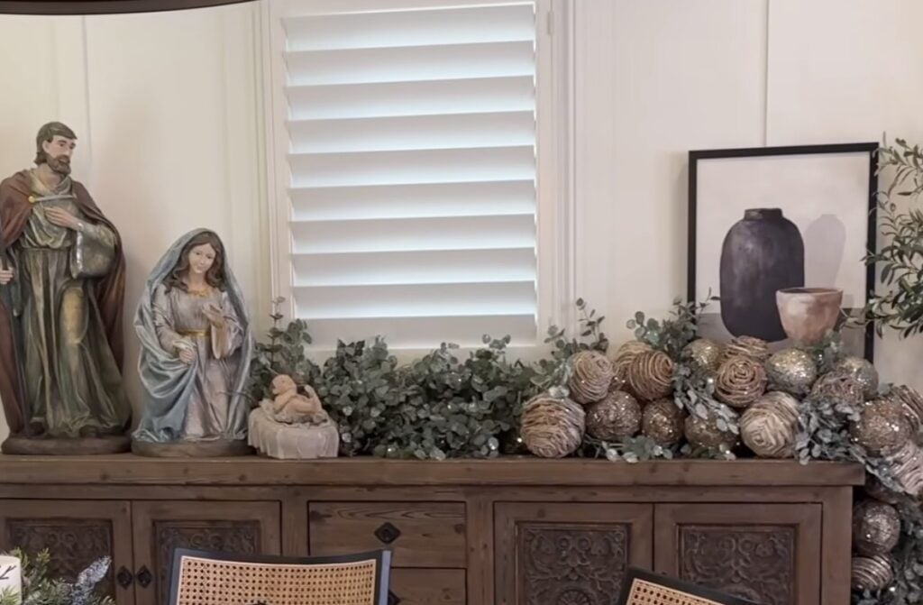 Mary, Joseph, and Baby Jesus displayed on a wooden sideboard with eucalyptus garland, oversized neutral ornaments, framed art, and a black vase.