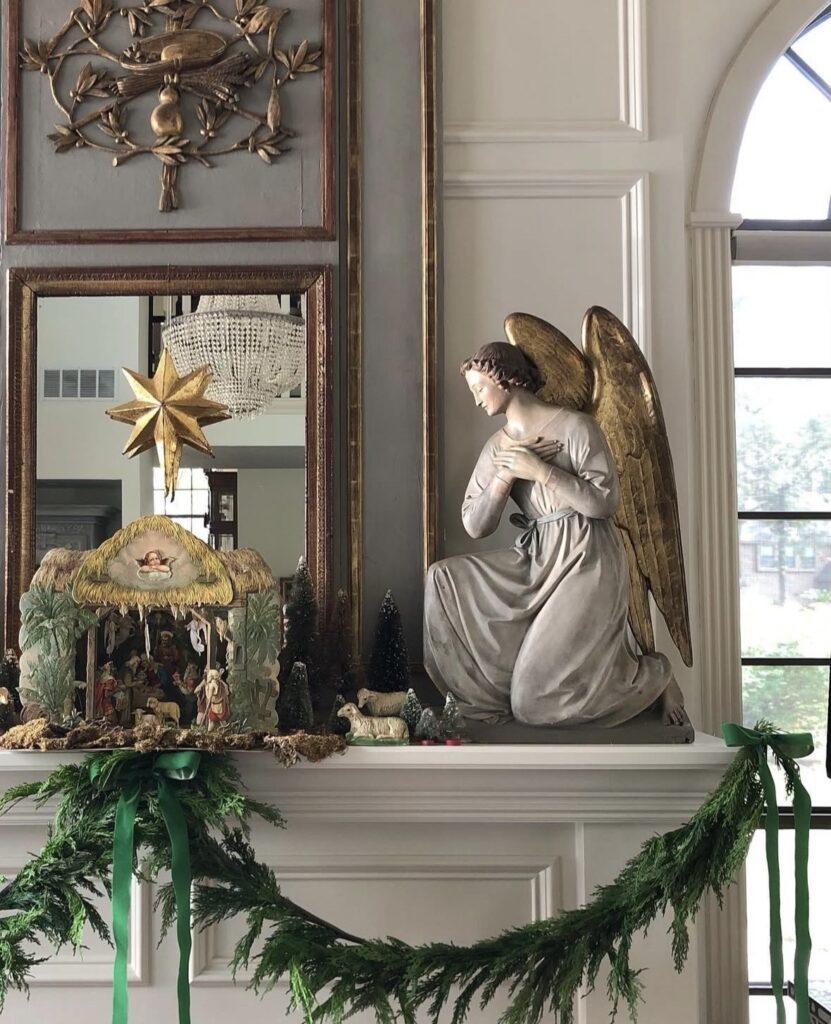 Traditional nativity scene and oversized kneeling angel displayed on a mantel with evergreen garland, green velvet bows, miniature trees, and a gold star.