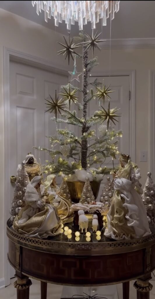 Gold and white nativity scene arranged on a round table around a slim Christmas tree decorated with metallic star ornaments and warm lights.