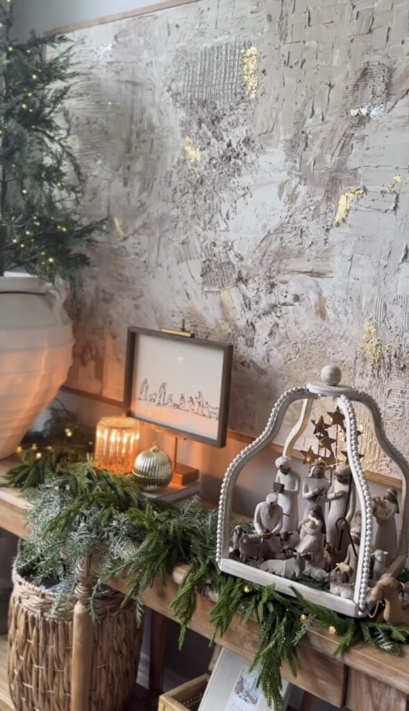 Neutral nativity scene displayed inside a beaded arched frame on a console layered with evergreen garland, candles, abstract artwork, and a small lit tree.
