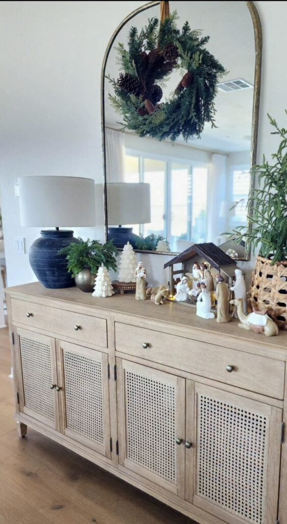 Neutral nativity scene styled on a light wood console with miniature white trees, greenery, a black lamp, and an arched mirror holding a Christmas wreath.