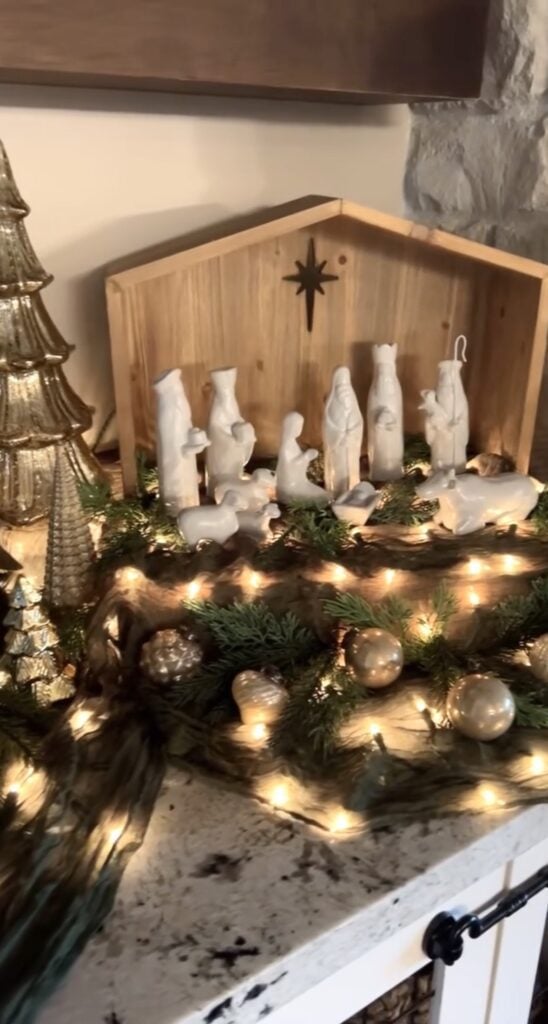 Modern white ceramic nativity figures displayed inside a simple wooden stable with evergreen garland, gold ornaments, metallic trees, and warm fairy lights.