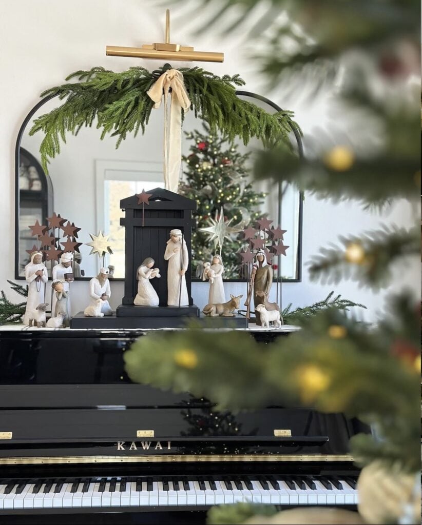 Willow-style nativity set arranged on a black piano beneath an arched mirror decorated with evergreen garland and a cream velvet bow.