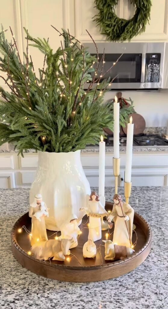Neutral nativity figures arranged on a round wooden tray with warm fairy lights, white taper candles, and a large vase filled with fresh evergreen branches.