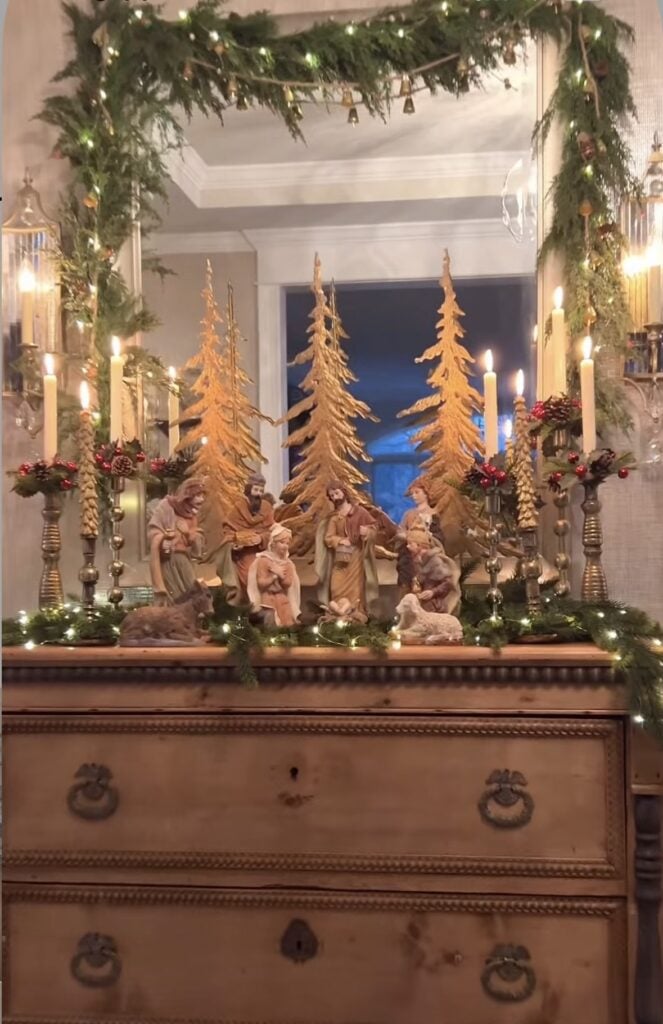 Traditional nativity figures displayed on a wooden dresser with tall carved Christmas trees, candlelight, red berries, and evergreen garland framing a mirror.