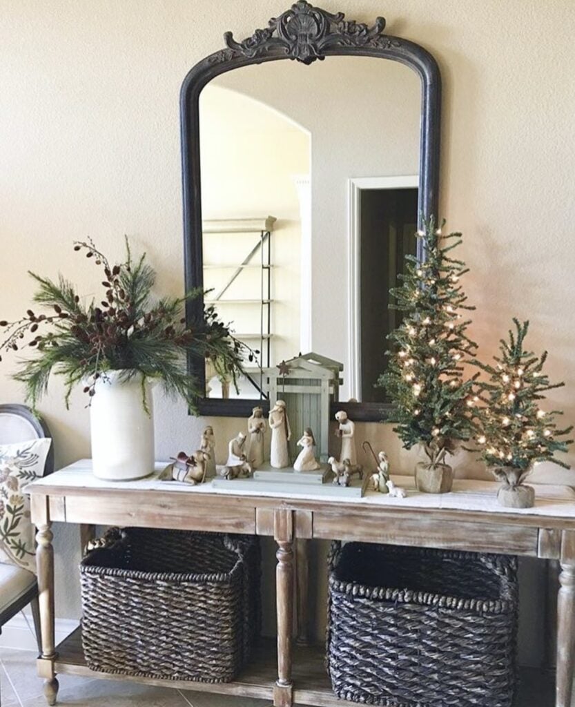 Neutral nativity set styled on a rustic console table with a large white vase of greenery, two lit tabletop trees, and a black framed mirror.