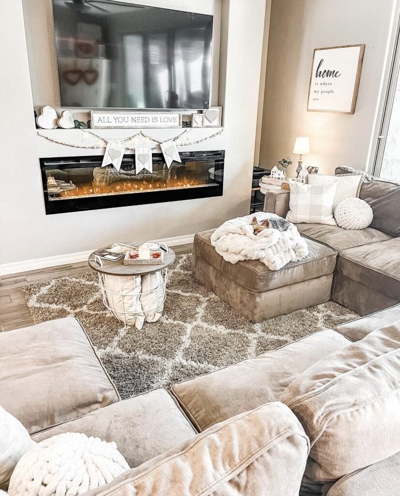 Cozy living room with a modern fireplace, “All You Need Is Love” sign, heart banner garland, and soft neutral pillows.