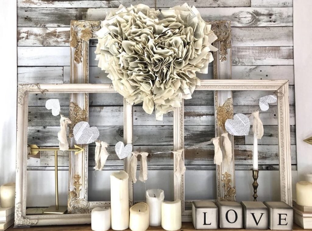 Neutral valentines decor ideas: Mantel shelf with distressed frames, a book-page heart wreath, white candles, and LOVE letter blocks in a neutral palette.