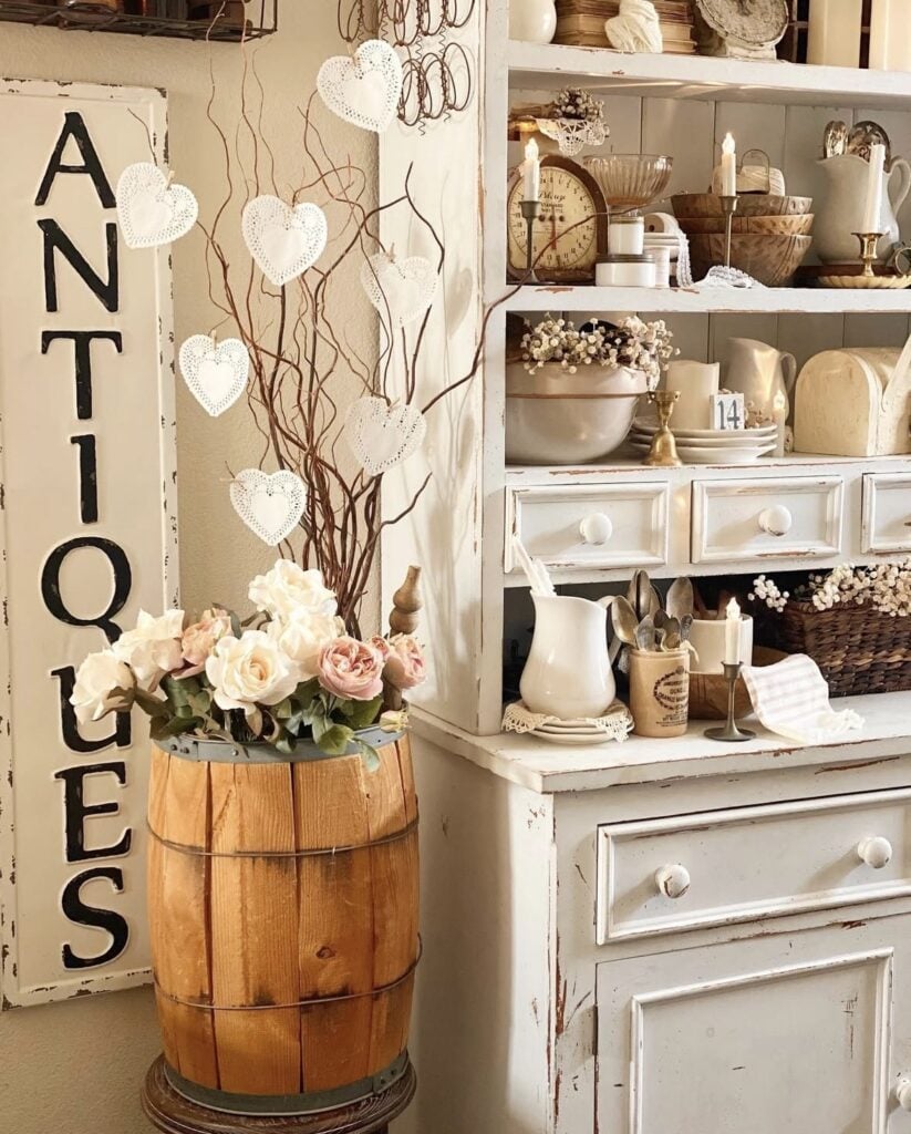 Vintage hutch corner with an “ANTIQUES” sign, lace hearts on branches, and roses in a wooden barrel for neutral Valentine decor.