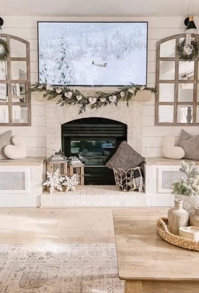 Cozy living room with a white fireplace, greenery garland with wooden hearts, lanterns, knit pillows, and snowy winter art on the TV.