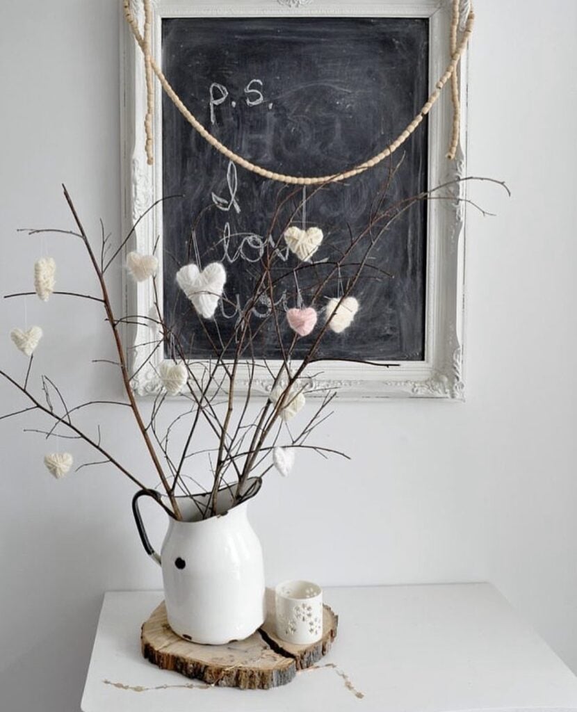 Neutral Valentine decor with bare branches in a white pitcher and tiny crochet hearts, styled under a framed chalkboard and wood bead garland.