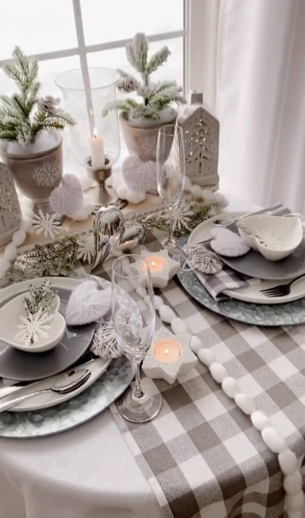 Neutral winter table setting with gray buffalo check runner, white heart ornaments, candleholders, pine greenery, and layered plates and glassware.