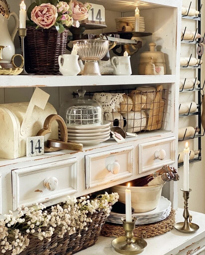 Vintage white hutch styled with stacked plates, glass cloche, baskets, soft florals, and warm candlelight for cozy neutral Valentine decor.