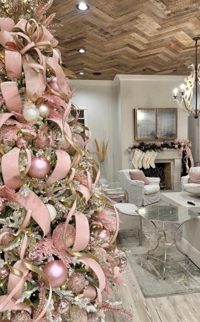 Close view of a Christmas tree layered with wide blush pink ribbon, gold ribbon, oversized pink ornaments, and sparkling floral picks.