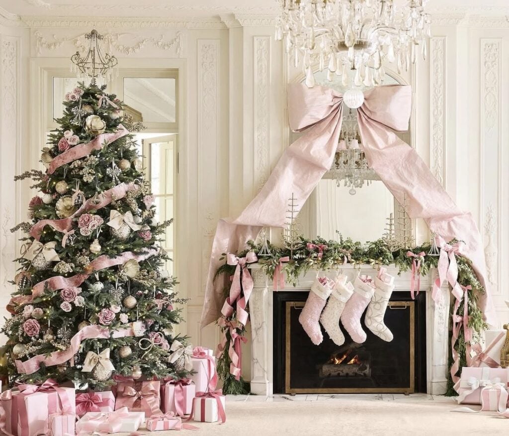 Elegant Christmas tree decorated with dusty pink ribbon, blush roses, and gold ornaments beside a fireplace draped with an oversized pink bow.