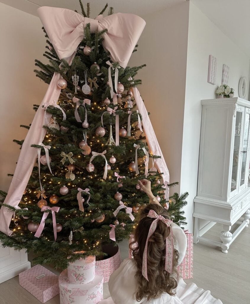 Green Christmas tree decorated with pale pink bows, blush ornaments, delicate hanging decorations, and an oversized pink bow topper.