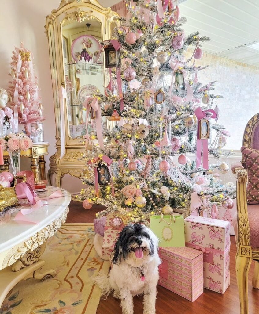 Flocked Christmas tree decorated with pink ribbons, blush ornaments, vintage-style framed decorations, and warm lights in an ornate pink and gold room.