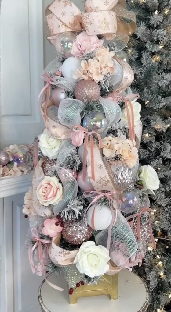 Small flocked Christmas tree decorated with blush pink and silver ornaments, roses, ribbons, and sparkling mesh.