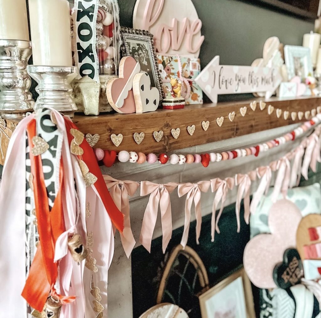 Pink Valentine’s Day mantel decorated with heart garlands, ribbon streamers, layered “love” signs, candles, and soft pink accents across a wooden mantle.