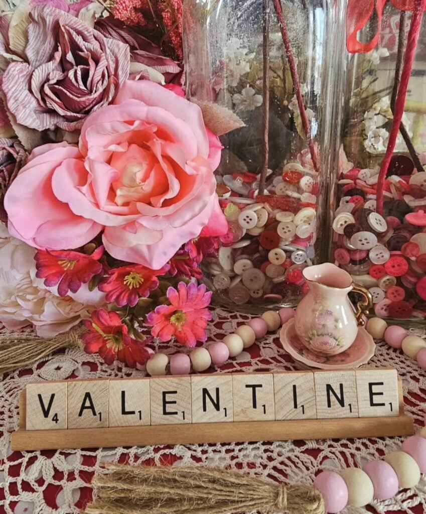 Vintage-inspired pink Valentine tabletop with layered faux flowers, a glass jar filled with pink and white buttons, bead garland, and a Scrabble tile “VALENTINE” sign.