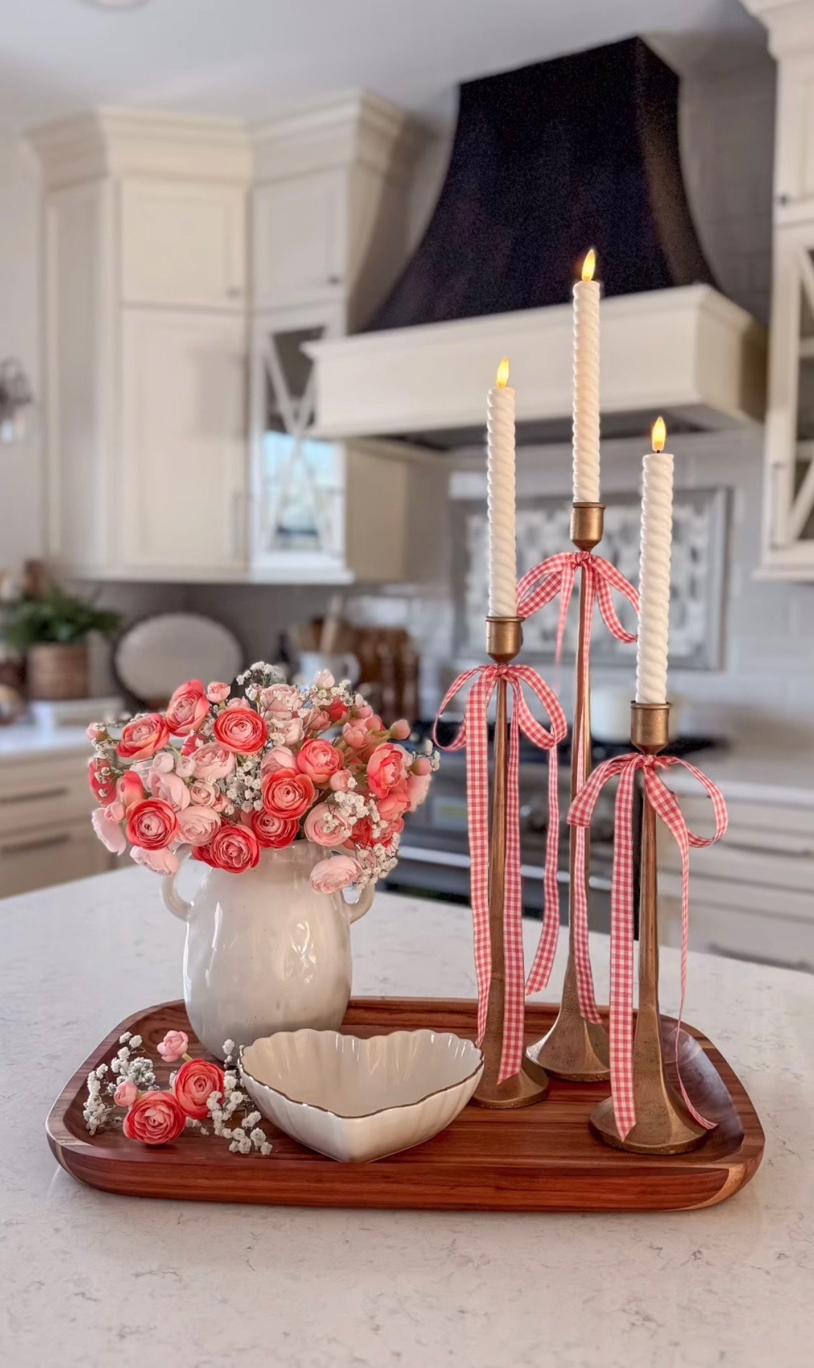 Soft pink Valentine kitchen island centerpiece with a blush rose bouquet in a white pitcher, three tall taper candles tied with pink gingham bows, and a heart-shaped bowl on a wood tray.