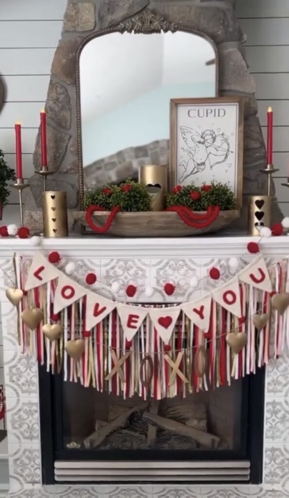 Valentine fireplace mantle with cupid print, red taper candles, and a “Love You” tassel banner.