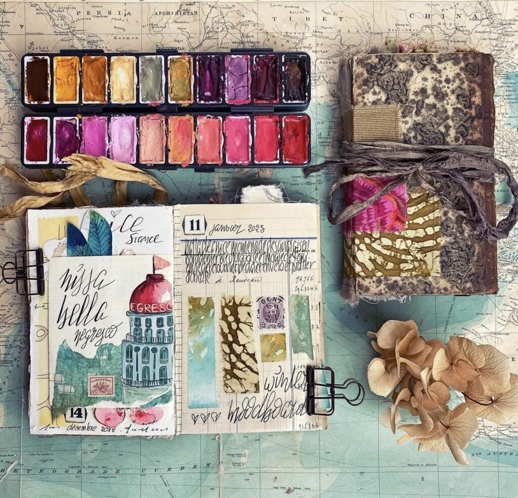 Flat lay of travel journaling supplies on a world map background, including watercolor palette, fabric-wrapped journal, mini sketchbooks, and dried flowers.