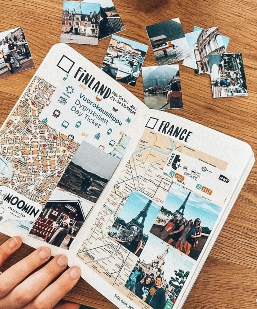 Small travel journal open to “Finland” and “France” pages with maps, ticket pieces, and photo collages, with printed travel photos scattered on a wooden table.