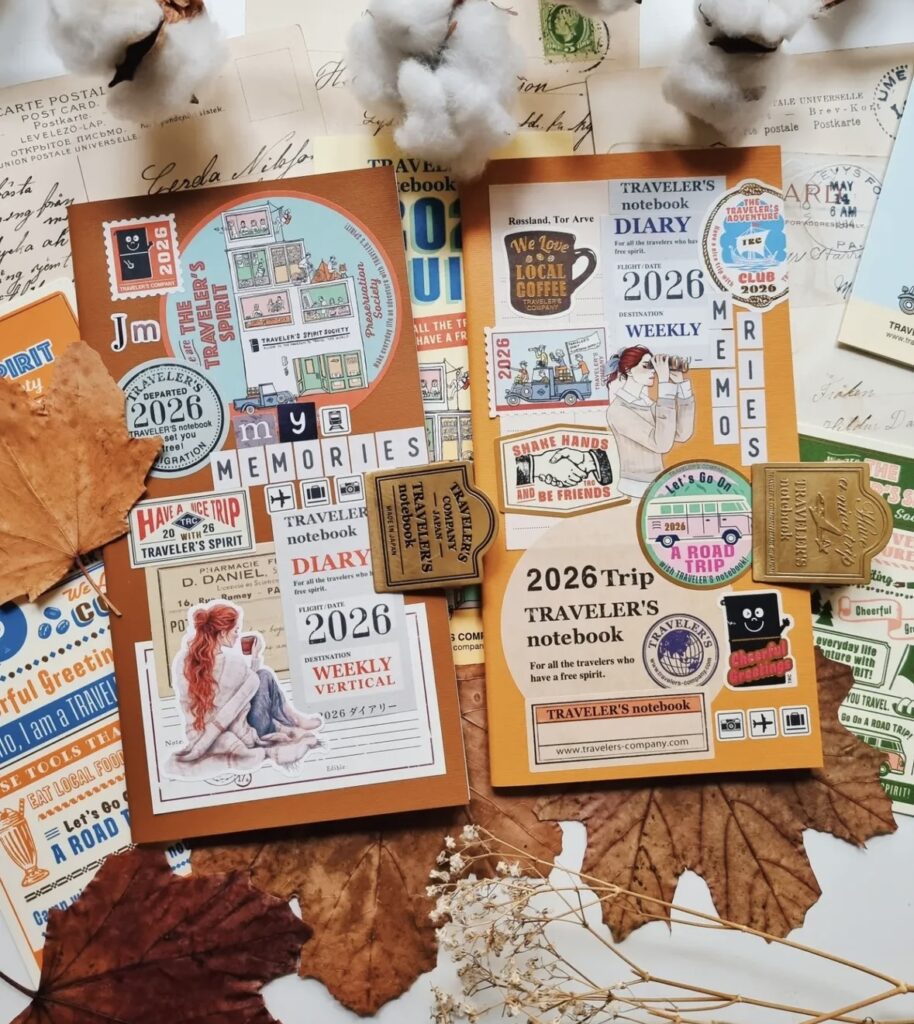 Flat lay of two Traveler’s Notebook 2026 diary covers decorated with stickers, stamps, “my memories,” “a road trip,” and travel icons, styled with autumn leaves and cotton stems.