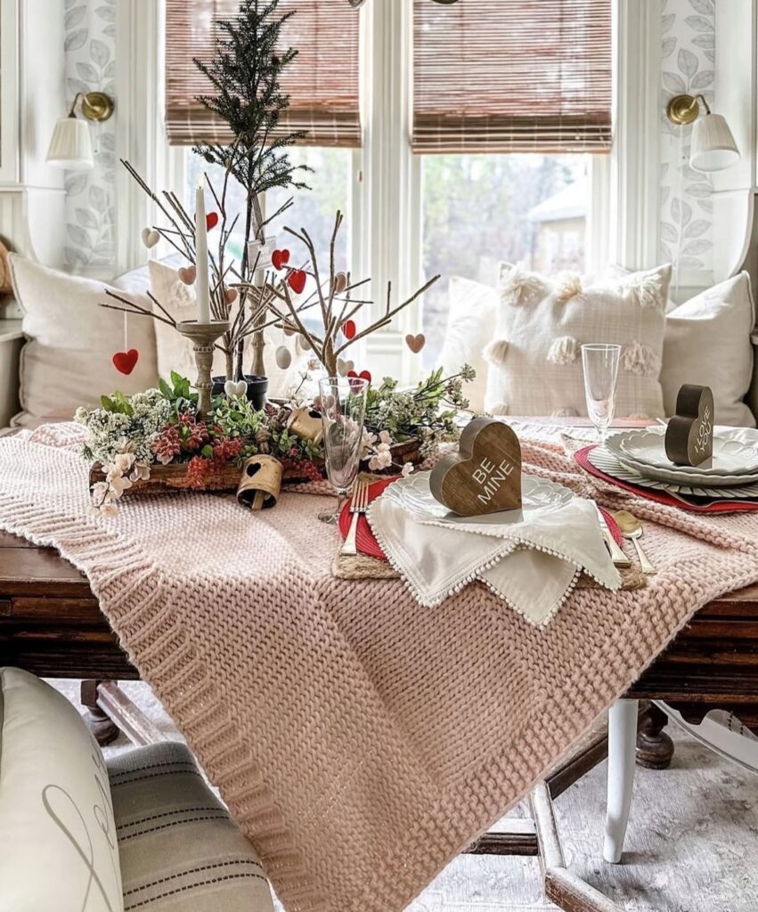 Cozy neutral Valentine tablescape with a blush knit runner, twig branches with red hearts, a wood candlestick centerpiece, and a “BE MINE” heart place card.
