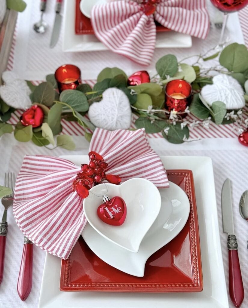 Valentine’s Day table setting with red striped napkin bow, heart-shaped plates, and red ornaments.