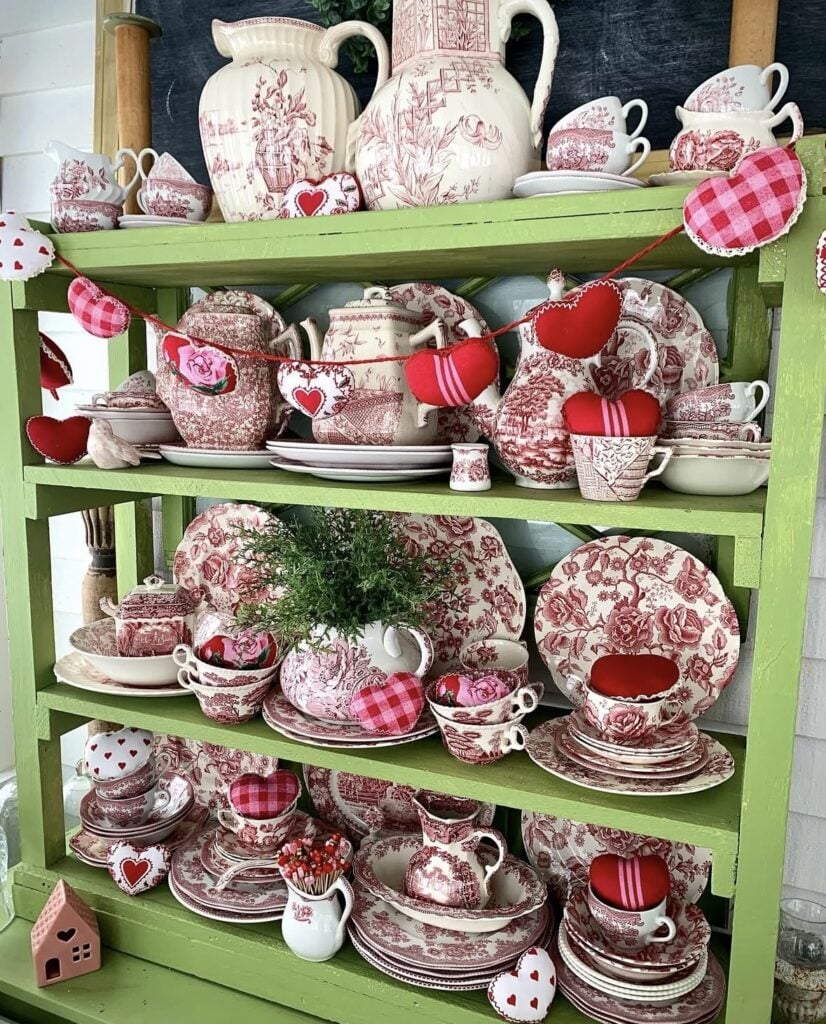 Green hutch Valentine decor with red toile dishes, stitched heart garland, and red and pink heart accents.