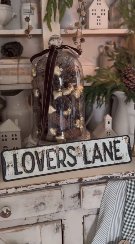 Rustic neutral Valentine’s shelf decor with a “Lovers Lane” sign and a glass cloche filled with pinecones and small gold hearts.