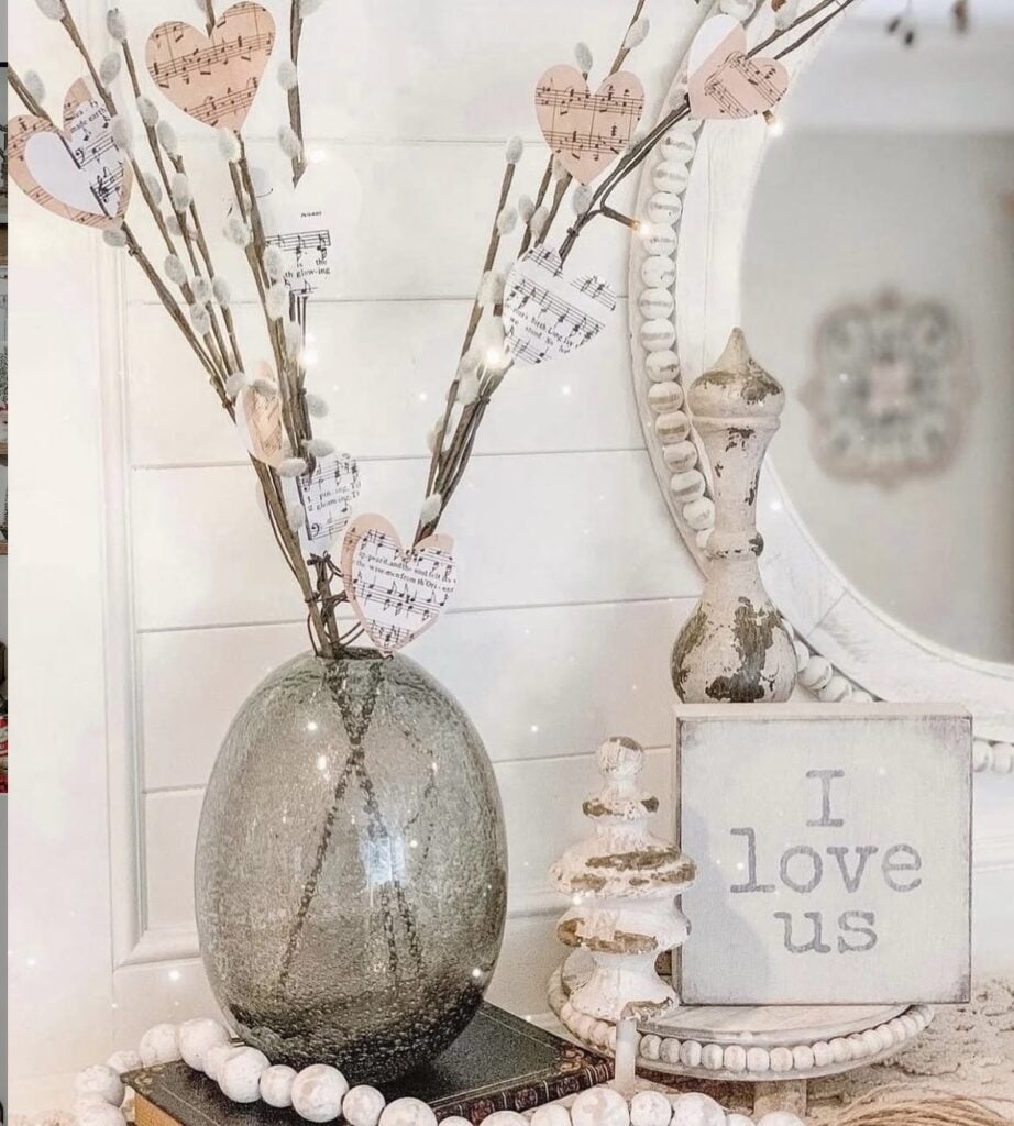 Neutral Valentine’s Day decor vignette with a crackled glass vase of pussy willow branches and sheet music paper hearts, plus an “I love us” sign and beaded garland.