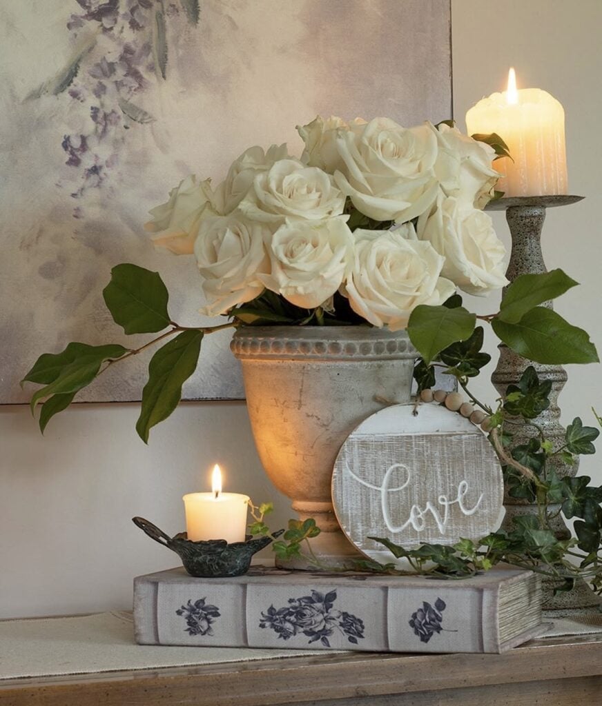 Neutral Valentine’s decor with white roses in a stone urn, candles, trailing ivy, and a round wood “Love” sign on a vintage floral book box.